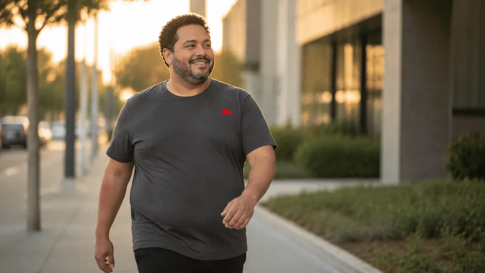 Homem plus size vestindo camiseta cinza chumbo lisa com leão vermelho pequeno no peito, caminhando na rua ao fim da tarde, luz dourada