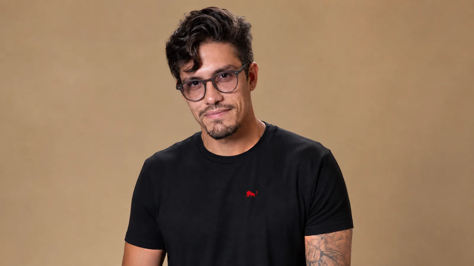 Pedro, fundador da Lion Brazil, em retrato editorial vestindo camiseta preta tecnológica de caimento impecável com o leão vermelho pequeno no peito esquerdo, fundo em tom areia