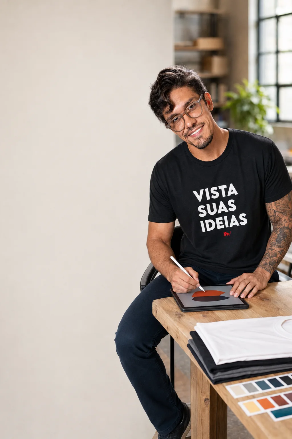 Cliente desenhando a própria arte num tablet, vestindo camiseta preta Vista suas ideias da Lion Brazil, com peças dobradas e cartela de cores na mesa