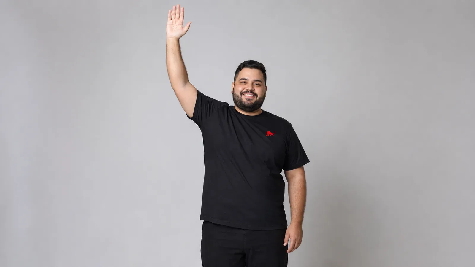 Homem plus size de barba vestindo camiseta preta lisa com leão vermelho pequeno no peito, em pé, luz natural lateral de estúdio