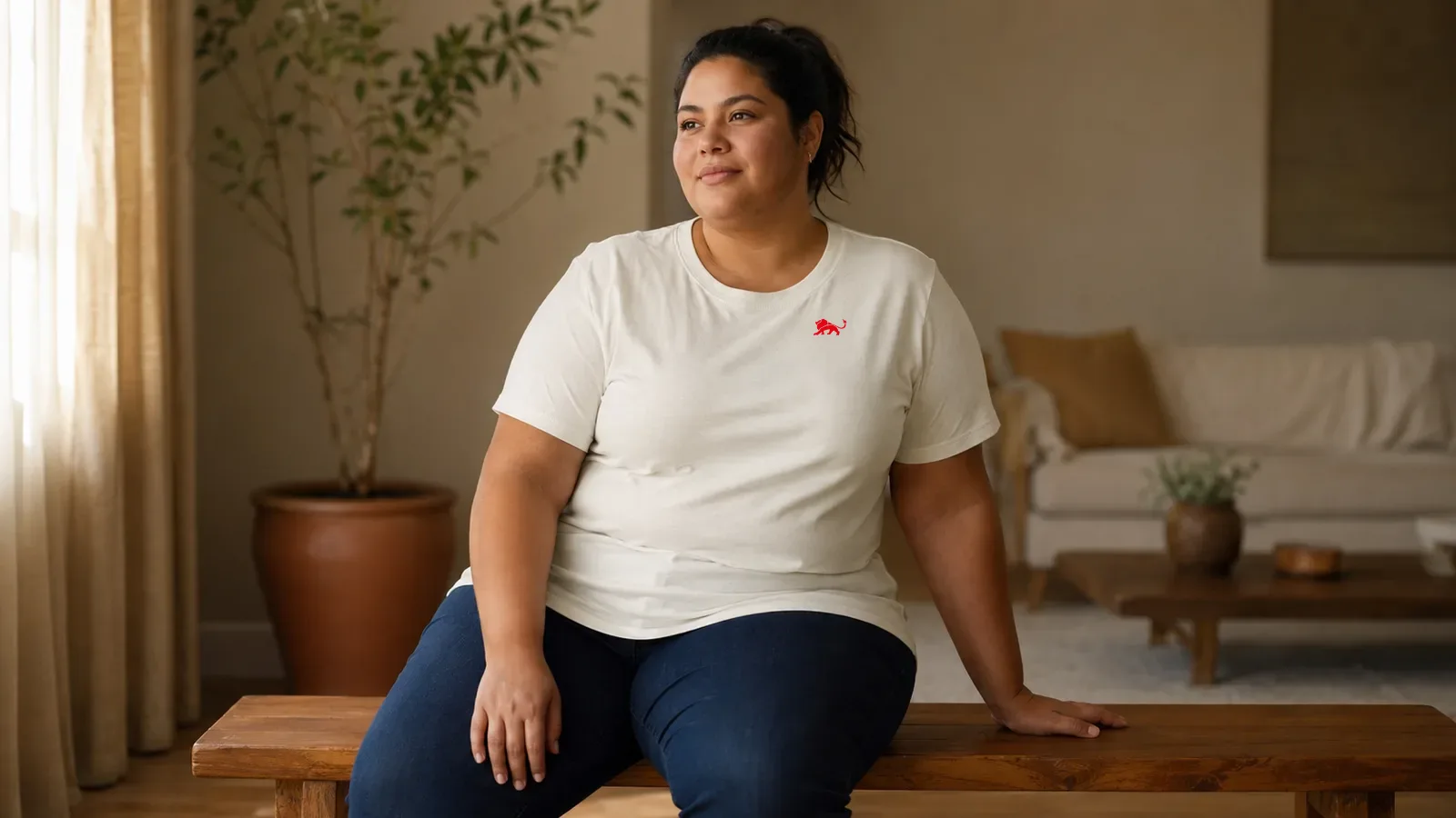 Mulher plus size de cabelo preso vestindo camiseta off-white lisa com leão vermelho pequeno no peito, sentada em banco de madeira, luz de janela