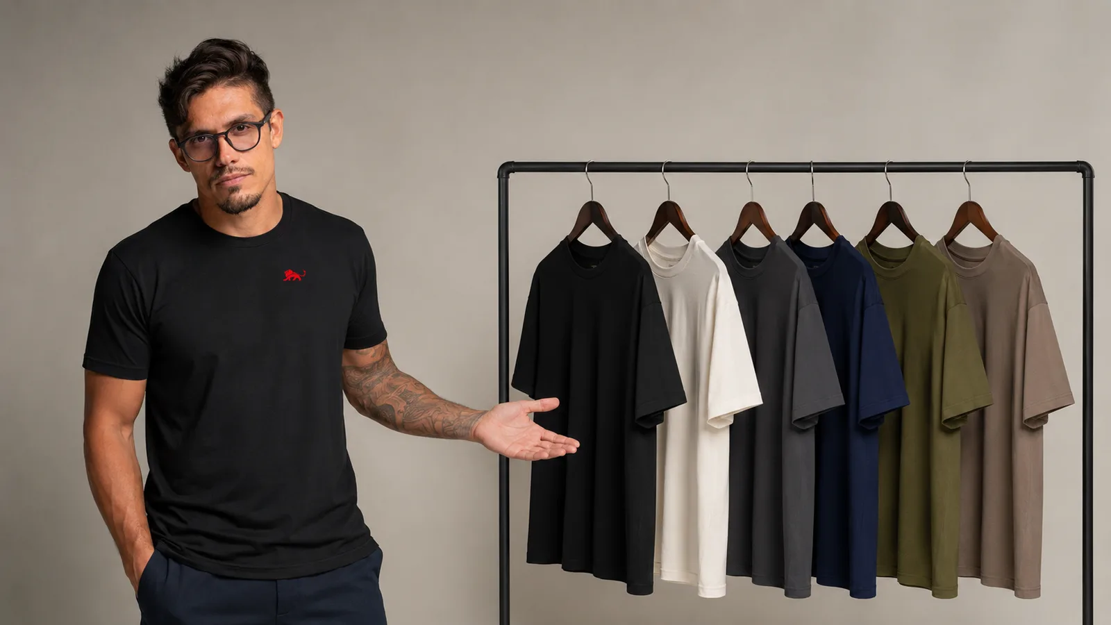 Pedro ao lado de uma arara com seis camisetas plus size lisas em seis cores sóbrias, vestindo camiseta preta com o leão vermelho pequeno no peito, fundo neutro de estúdio