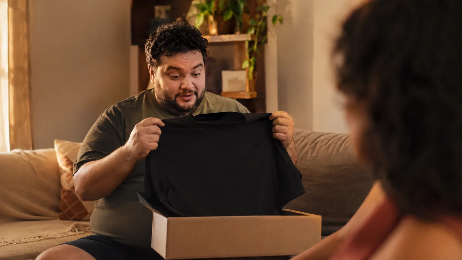 Homem plus size abrindo uma caixa de presente e segurando uma camiseta preta com leão vermelho pequeno no peito, sala de estar, luz de fim de tarde