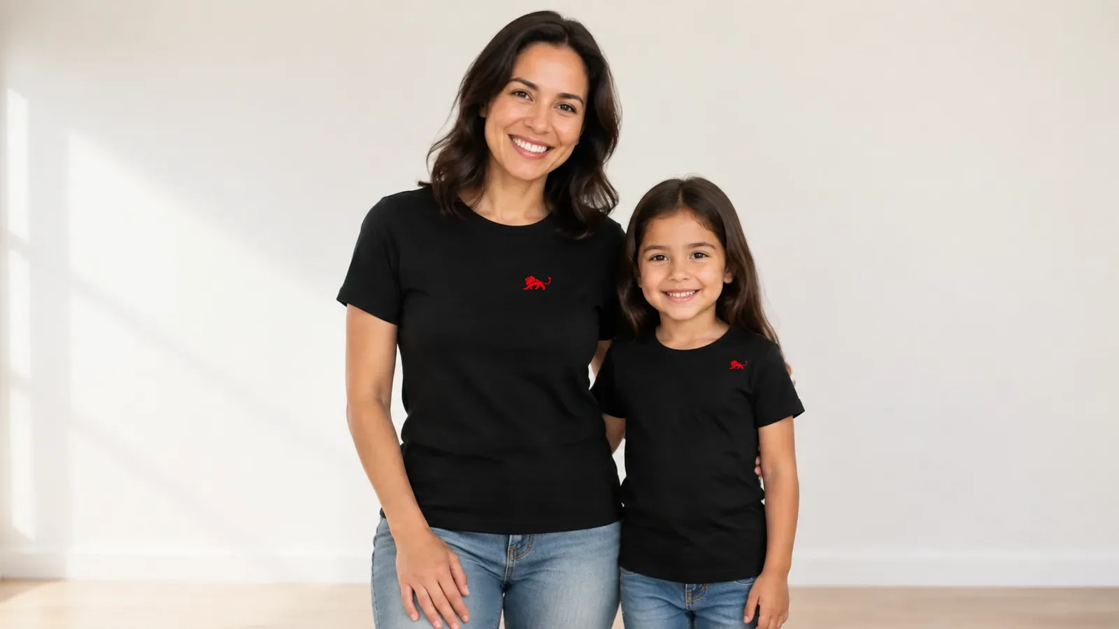 Mãe e filha de uns 6 anos lado a lado, de frente, com a mesma camiseta preta e a arte ajustada em cada tamanho, o leão vermelho pequeno no peito, fundo claro