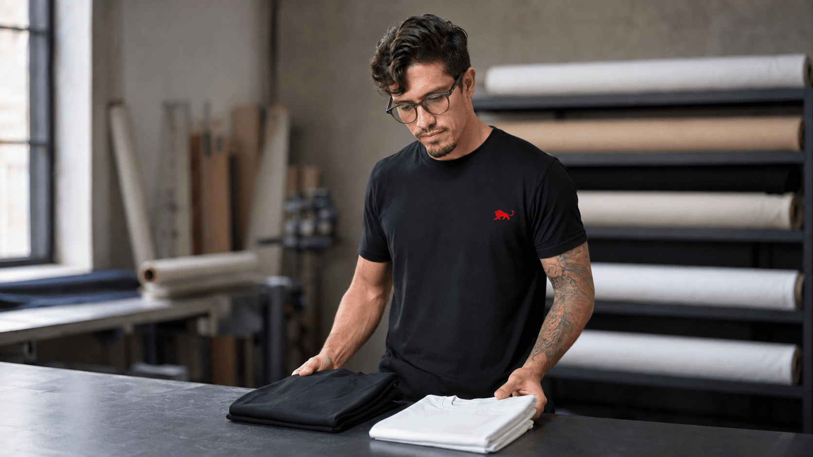 Pedro, fundador da Lion Brazil, na bancada do ateliê examinando duas camisetas dobradas, uma preta e uma branca, vestindo camiseta preta com o leão vermelho pequeno no peito, rolos de tecido ao fundo